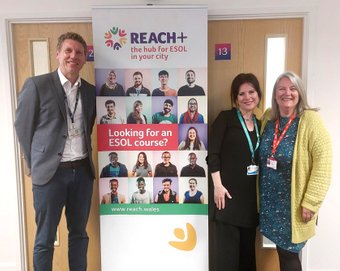 Three individuals standing in front of a banner promoting ESOL courses offered by REACH+
